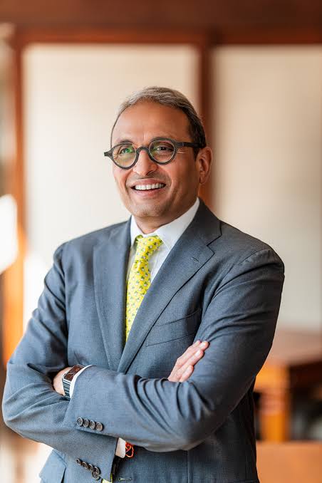 Russell Mehta standing with his arms folded, in a tailored grey suit and yellow tie, in a wood-panelled room.