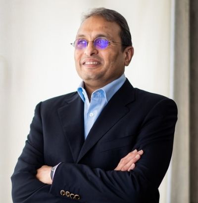 Russell Mehta in a navy blazer over an open-collar blue shirt, arms folded, looking away from the camera.