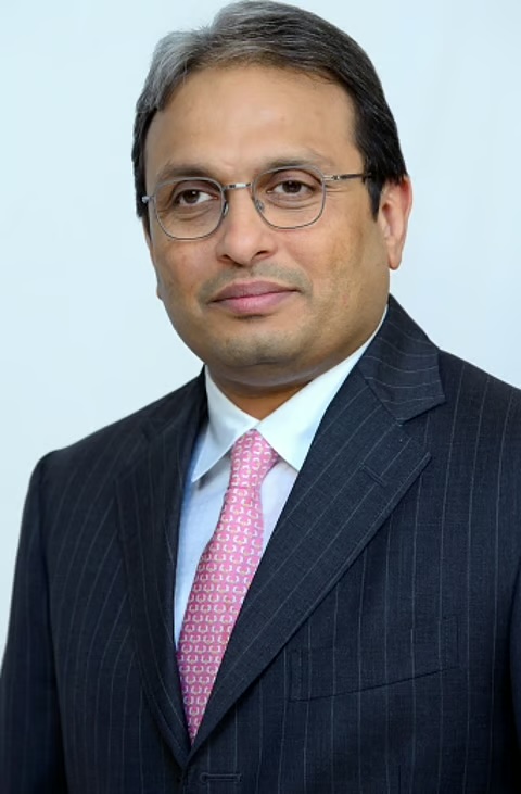 Formal portrait of Russell Mehta in a dark pinstriped suit and pink patterned tie, against a pale background.