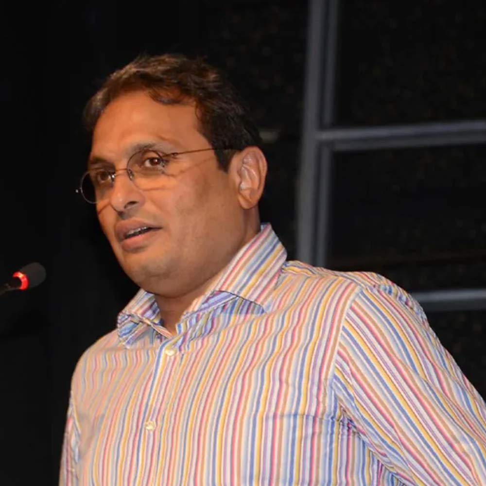Russell Mehta speaking at a microphone in a striped shirt during an earlier industry address.